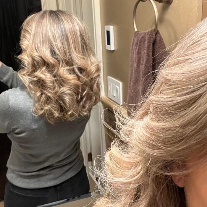 A person, seen from the back, stands in front of a mirror showcasing curly, shoulder-length hair in a bathroom. Only the hair and a partial view of the head are visible.