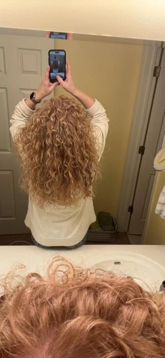 A person with curly hair takes a mirror selfie, capturing the back of their head. The bathroom, including a sink and a door, is visible in the reflection.