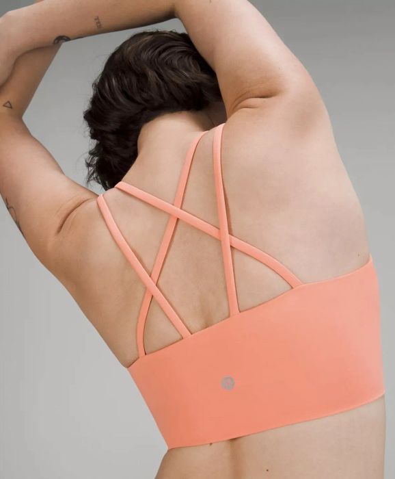 Model stretching her arms upward and to the side, showcasing the strappy back of the peach sports bra.