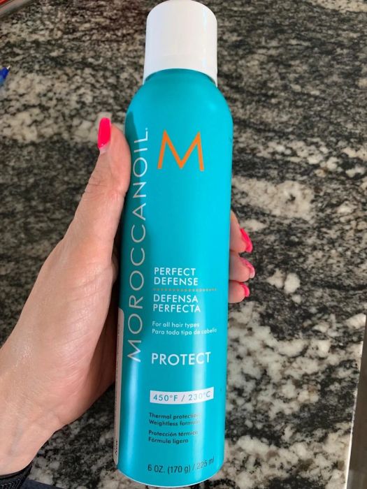 A hand holds a bottle of Moroccanoil Perfect Defense, a thermal protection spray suitable for all hair types, featuring product details in both English and Spanish.
