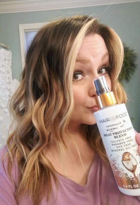 A woman is holding a bottle of Hair Food Coconut & Argan Oil Heat Protection Blend Spray, smiling gently. The product is clearly displayed in her hand.