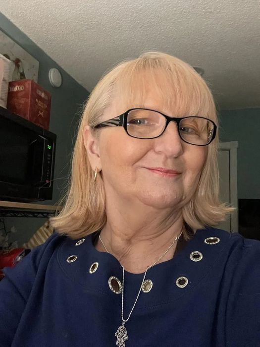 A woman with short blonde hair and glasses, wearing a round-neck top with metallic embellishments and a necklace with a hand-shaped pendant.