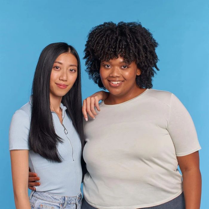Two women stand together, one with long straight hair wearing a polo shirt and the other with short curly hair in a short-sleeve top, both smiling against a plain background.