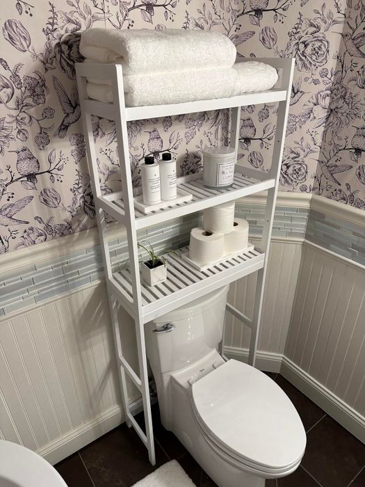 Bathroom shelving unit placed above the toilet with towels, toiletries, and toilet paper neatly arranged on the shelves