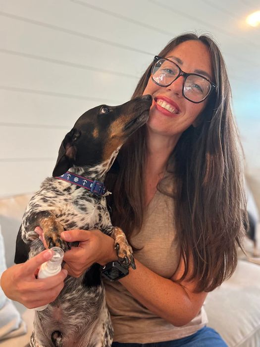 A woman with glasses smiling while cuddling a small dog that is licking her face, both indoors on a couch
