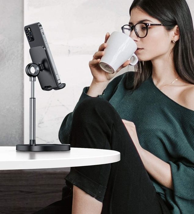 A model watching something on their phone, which is propped up at eye level on a black stand placed on a desk.