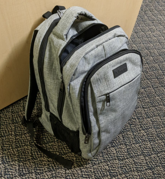 A review image showing a gray backpack standing upright on the floor, displaying multiple front pockets, a laptop sleeve, and ample storage.