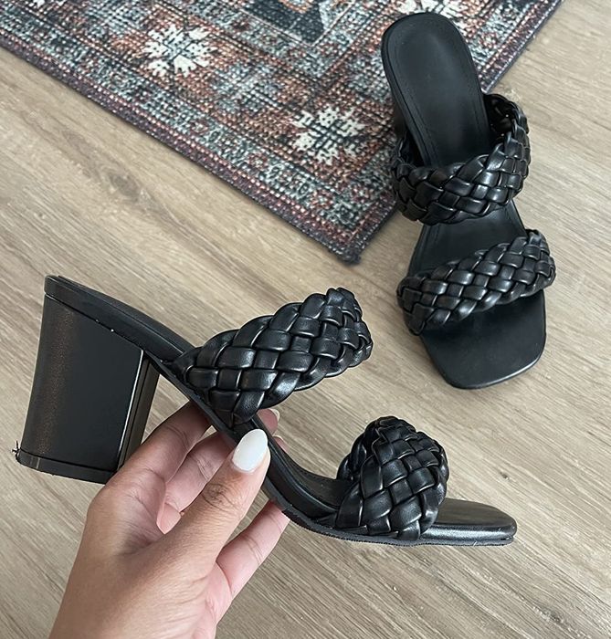 An individual holding a braided black heeled sandal beside its matching pair on a wooden floor.