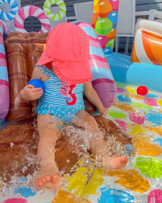 A baby, wearing a sun hat and a seahorse-themed swimsuit, happily splashes in a vibrant inflatable pool.