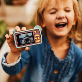 A child happily holds a toy camera, with their photo displayed on the screen. The child beams a wide smile, dressed in a casual outfit.