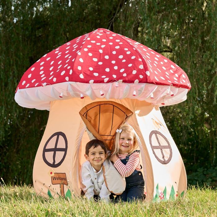 Two children grinning and peeking out from inside a mushroom-shaped play tent with charming window designs, outdoors. A 'Welcome' sign hangs above the door.