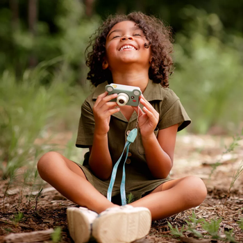 A young child, sitting cross-legged outdoors with a smile, holds a toy camera while wearing a casual, comfortable outfit, eager to capture the world around them.