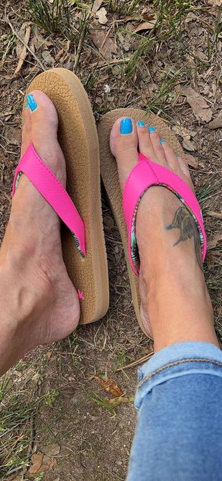A person wearing bright pink flip-flops with a bird tattoo on their foot, standing on the ground beside a patch of grass.