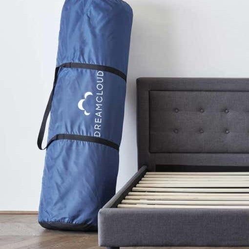An empty bed frame positioned next to a large DreamCloud mattress stored in a canvas bag.