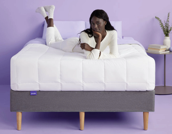 A model resting on a Purple mattress, featuring a white mattress cover and purple walls in the background.