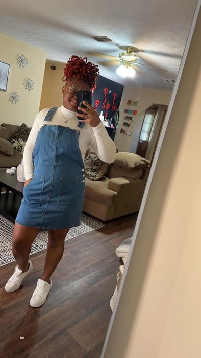 A reviewer taking a mirror selfie, wearing a denim overall dress paired with a white long-sleeve mock-neck bodysuit, accessorized with a headwrap and white sneakers.