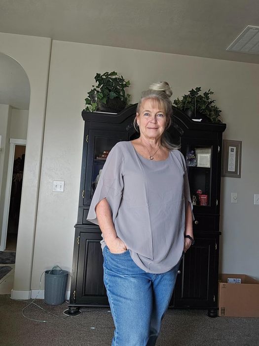 A woman in a casual grey top and blue jeans, striking a pose with hands on hips in a room.