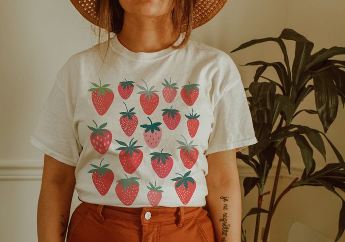 A model wears a white T-shirt featuring a strawberry print, paired with brown trousers and a straw hat, standing indoors near a plant.