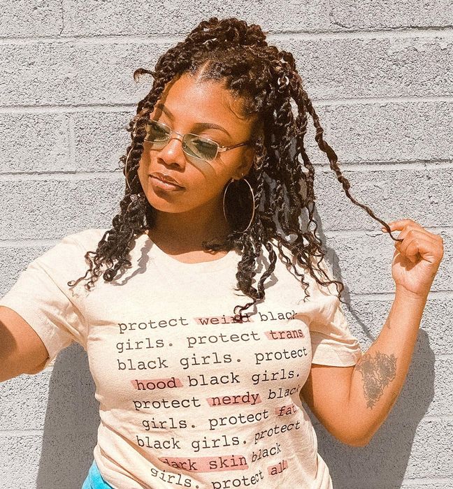 A model showcases a 'Protect Black Women' tee with a powerful message: 'Protect weird black girls, protect trans black girls, protect hood black girls, protect nerdy black girls, protect fat black girls, protect dark skin black girls, protect all black girls.'