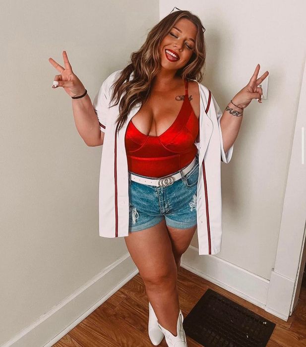 A woman in a red underwire bodysuit, denim shorts, and white boots poses while flashing peace signs.