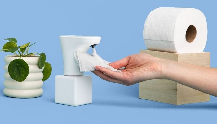 A hand holding toilet paper with foam on it, positioned under the Fohm dispenser, next to a roll of toilet paper and a potted plant.