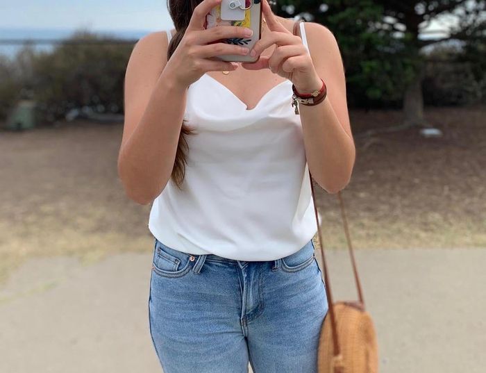 Reviewer posing for a selfie in a white satin cowl neck top and denim jeans, holding a straw bag.