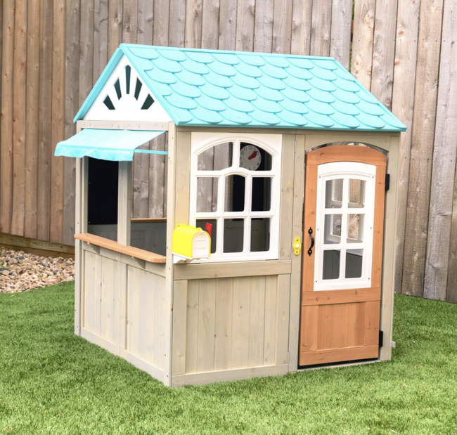 A charming playhouse sized for children, featuring a light blue roof with tiles, white-framed windows, a dark wooden door with glass panes, and walls resembling tan wood.