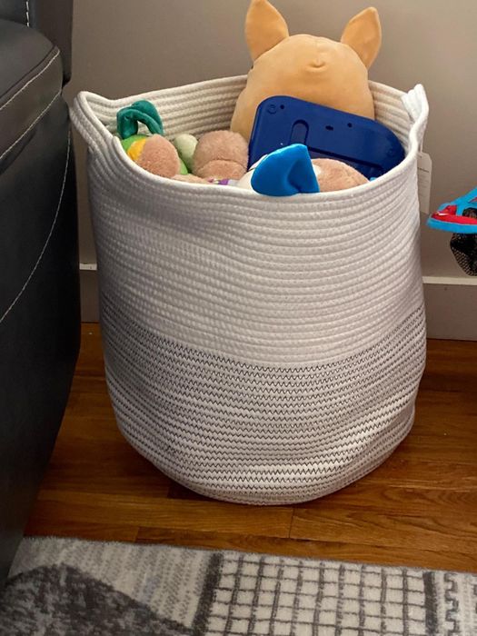 A woven basket filled with plush toys and a small blue tablet, placed next to a couch on a wooden floor