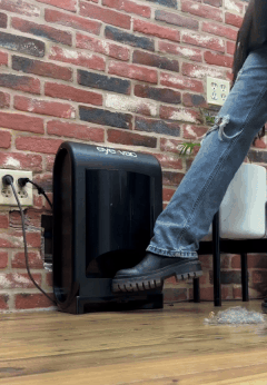 A person using a foot pedal to open a trash can placed against a brick wall.
