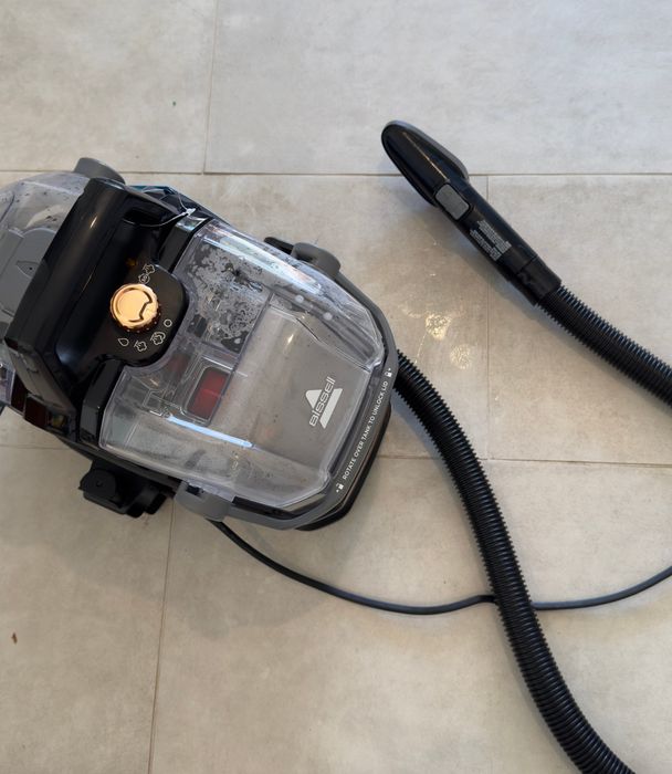 A carpet cleaning machine with a transparent canister and attached hose resting on a tiled floor.