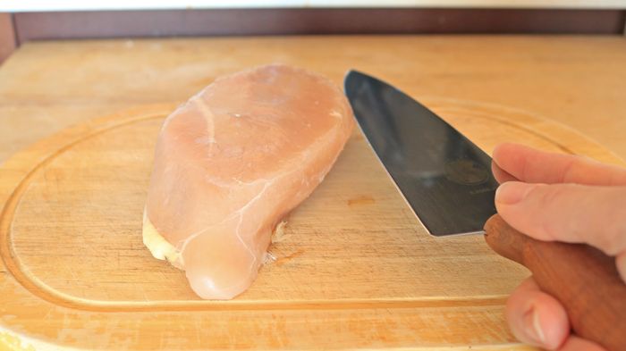 A knife positioned horizontally across a chicken breast on a cutting board.