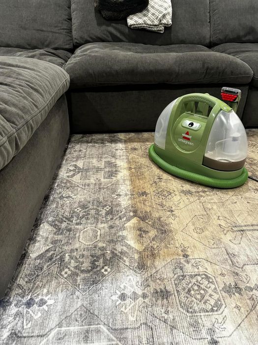 A Bissell Little Green machine is in action, cleaning a stained carpet next to a sectional sofa.