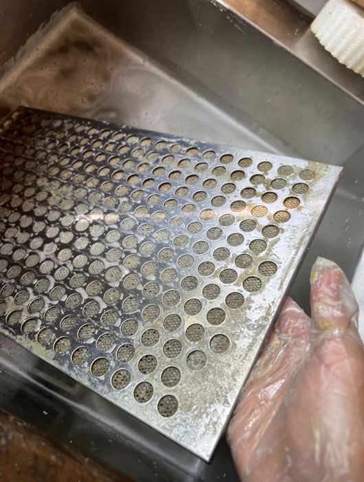A person wearing disposable gloves cleans a perforated metal tray in a sink full of soapy water.