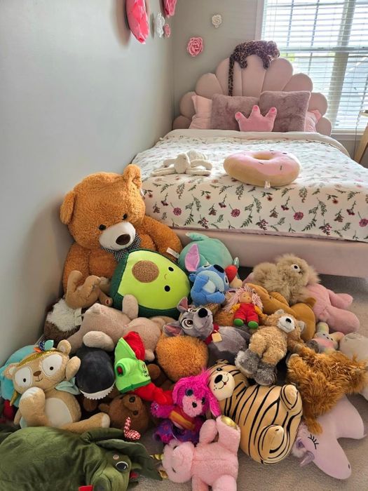 A cozy and playful bedroom setup with a variety of stuffed animals arranged on the floor near a child's bed, complete with a crown-shaped pillow for extra charm.