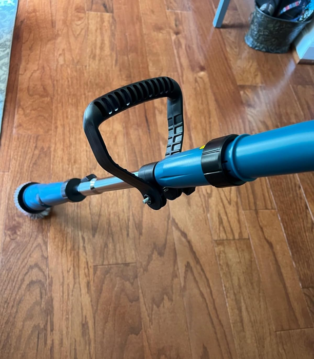 a close-up shot from the reviewer showing a scrubber tool with a black handle and blue shaft resting on a wooden floor