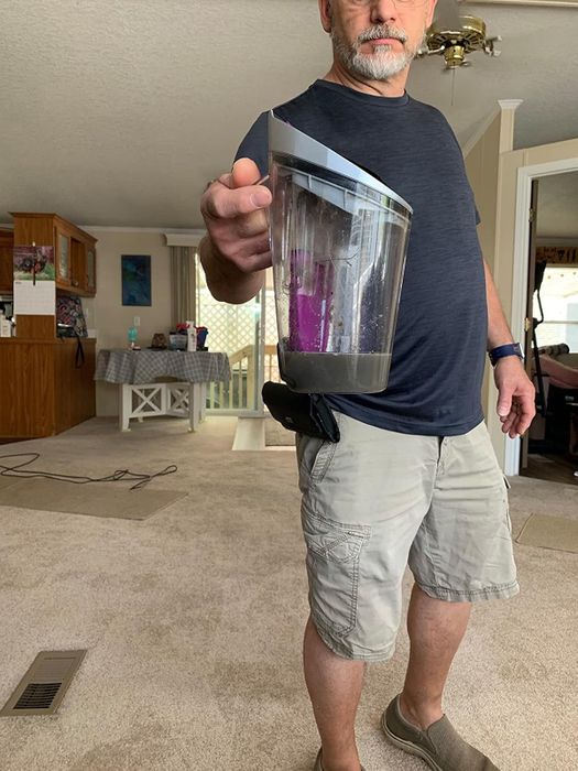 A man in casual clothing holding a vacuum dust container in a living room setting.