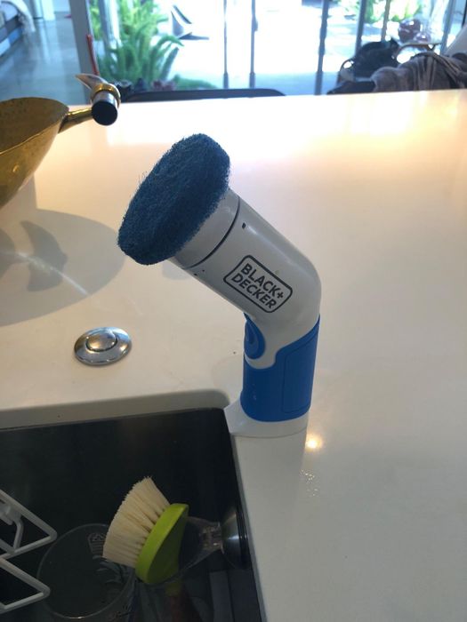 A Black & Decker power scrubber resting on a kitchen counter next to a sink, accompanied by a dish brush.