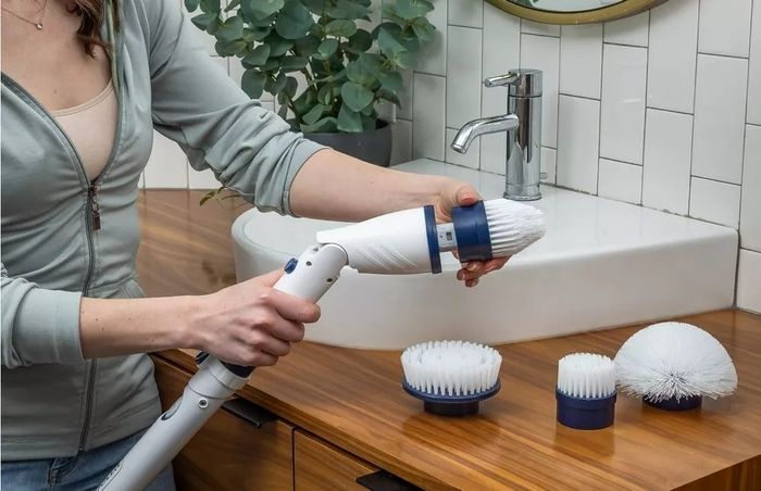 A person in a casual outfit is demonstrating a cleaning tool with multiple bristle head attachments in a bathroom environment. Text: