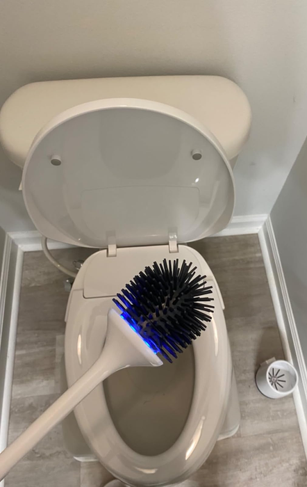 A reviewer is seen holding an electric silicone toilet brush with a blue light illuminated above their open toilet bowl.