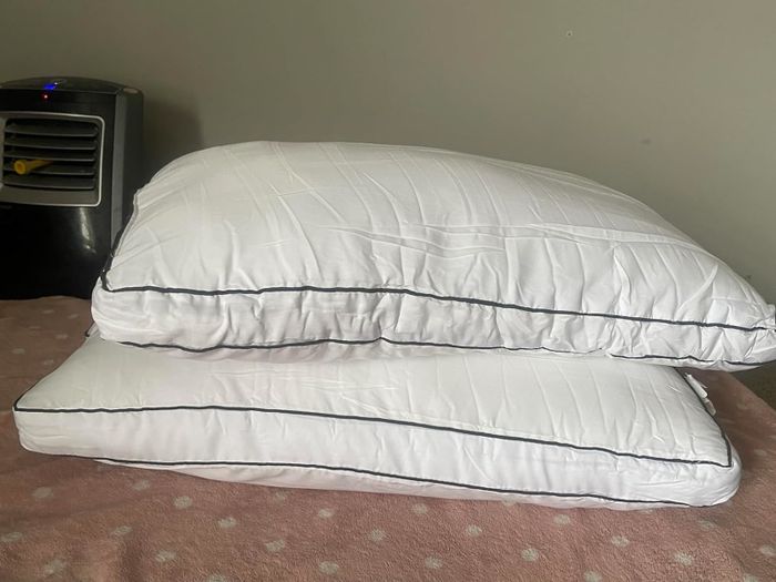 Two white pillows with black piping are stacked on a pink blanket, with a portable air conditioner visible in the background