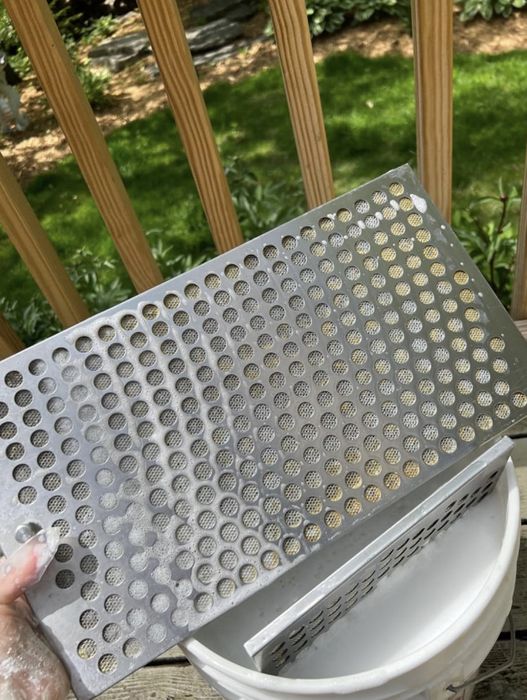 A hand gripping a large metal grate with perforated holes positioned above a white bucket. Behind this, a wooden fence and lush greenery are visible.