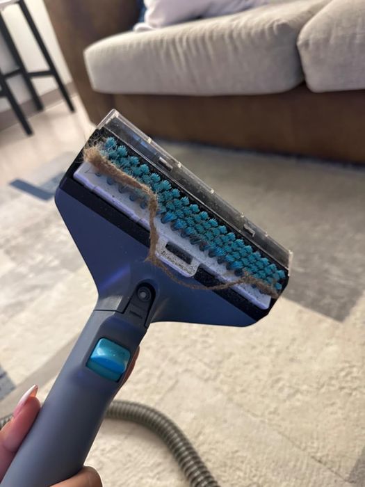 A hand holding a vacuum attachment with hair wrapped around the bristles, placed on a living room carpet.