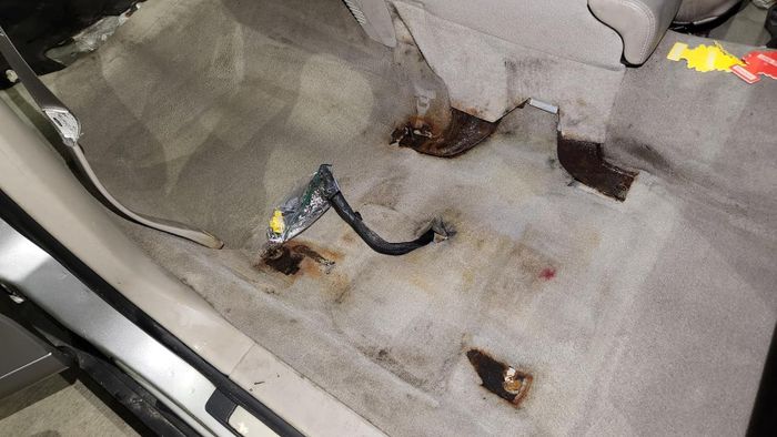 The inside of a car with the back seat removed, showing a floor stained and rusted. Wires and connectors are visible.