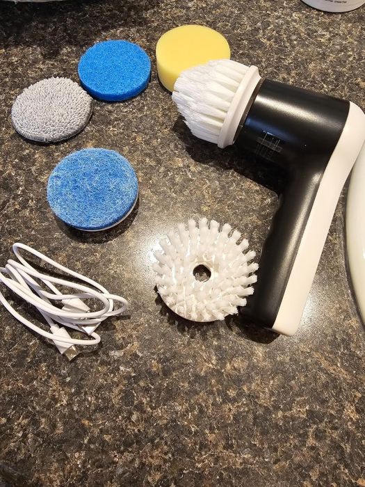 The reviewer’s electric cleaning brush with interchangeable scrub heads and a USB charging cable placed on a countertop.