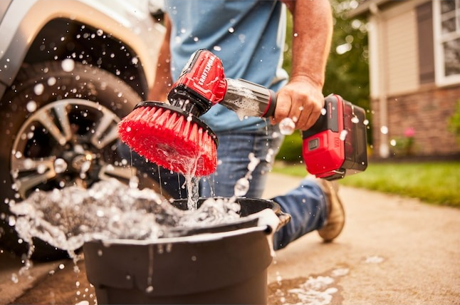 A person cleaning a car with a cordless power scrubber next to a bucket of water, causing splashes around them.