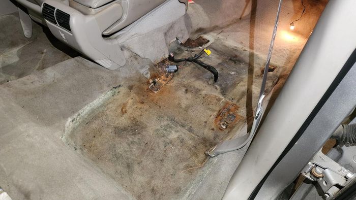 A car interior with the back seat removed, revealing exposed metal brackets, loose wires, and stained carpet flooring.