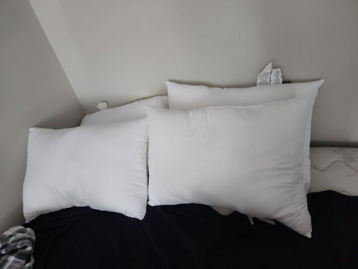 Three white pillows are neatly stacked against a wall on a bed adorned with a dark blanket. The tags on the pillows are clearly visible