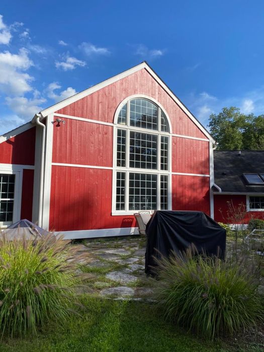 A charming red barn-style house with expansive arched windows and a patio adorned with covered outdoor furniture, surrounded by lush green plants in the foreground.