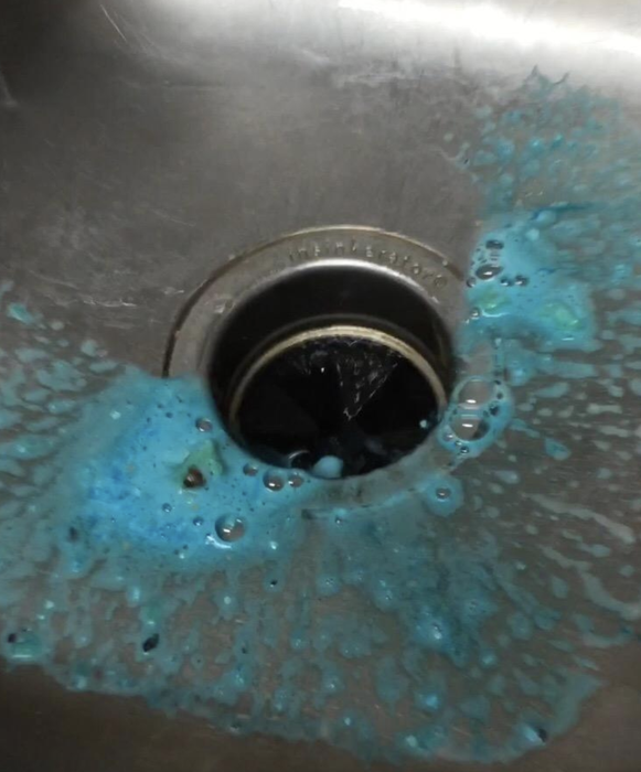 A sink drain surrounded by blue liquid, illustrating the use of the foaming cleaner.
