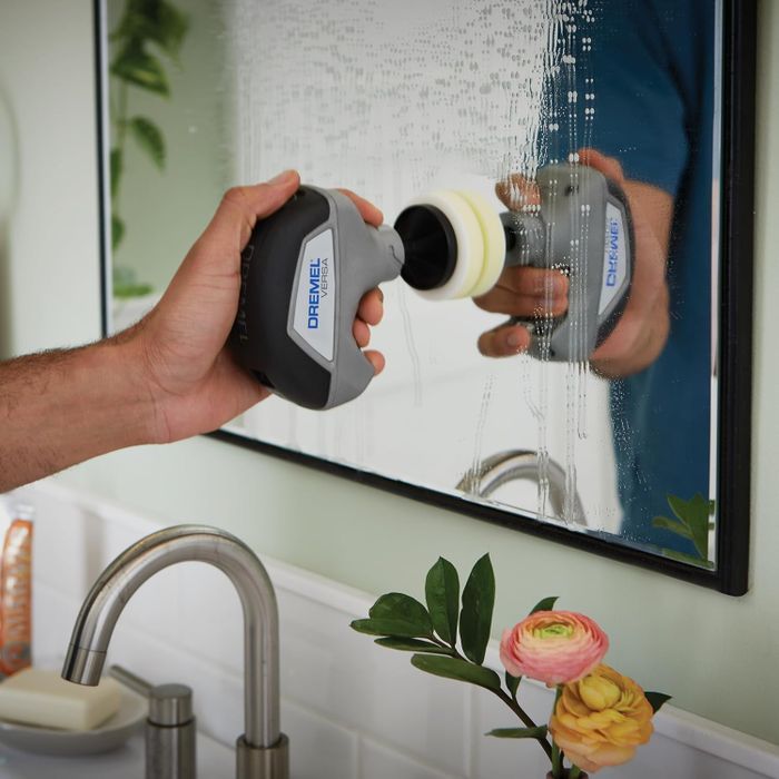 A hand is using the Dremel power scrubber to clean a bathroom mirror above a sink, with flowers and cleaning supplies visible in the background.